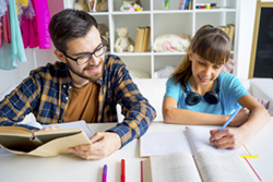 A father holding a textbook open while his teen daughter does schoolwork