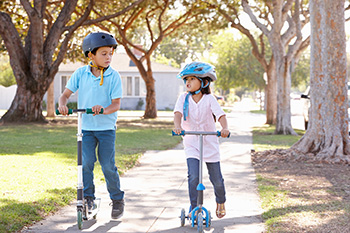 A boy and girl wearing helmets while riding scooters.
